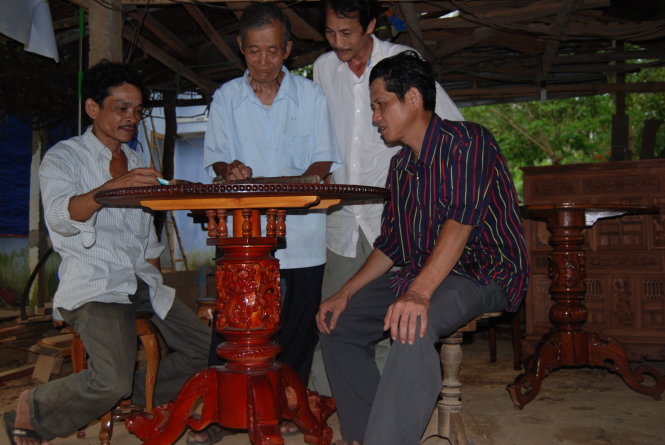 Ông Thẩm cùng với anh Phạm Miên (đứng kề ông Thẩm) và anh Trần Ngọc Tuấn (ngồi, bìa phải) ở tổ hợp tác mộc Văn Hà bàn cách làm đẹp cho những chiếc bàn xoay sắp đóng bằng cách thay đổi các họa tiết chạm khắc trên bàn - Ảnh: H.V.Mỹ