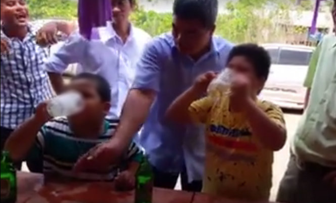 Người đàn ông áo xanh (giữa) liên tục ép 2 đứa trẻ uống liên tục các cốc bia -Ảnh cắt từ clip.