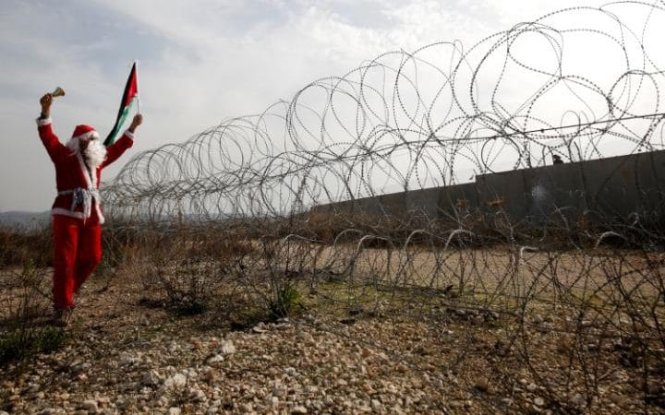 Một người biểu tình Palestine trong trang phục ông già Noel cầm lá cờ Palestine phản đối hàng rào của Israel ở làng Bilin tại khu vực Bờ Tây - Ảnh: Reuters
